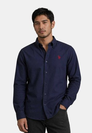 Man wearing dark blue button-up shirt with red embroidered logo, hand in pocket, standing against plain light background.