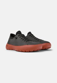Black synthetic shoes with a textured upper, flat laces, and a contrasting red rubber sole featuring grip patterns. Minimalist design.