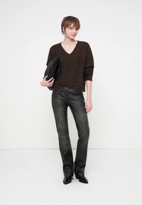 Junge Person mit kurzen Haaren, die einen braunen V-Ausschnitt-Pullover, schwarze Lederhosen, schwarze Stiefel trägt und eine schwarze Clutch-Tasche hält.