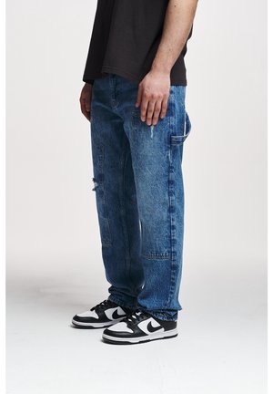 Mann trägt lockere blaue Jeans mit Seitentaschen und schwarz-weißen Nike-Sneakers, steht vor weißem Hintergrund.