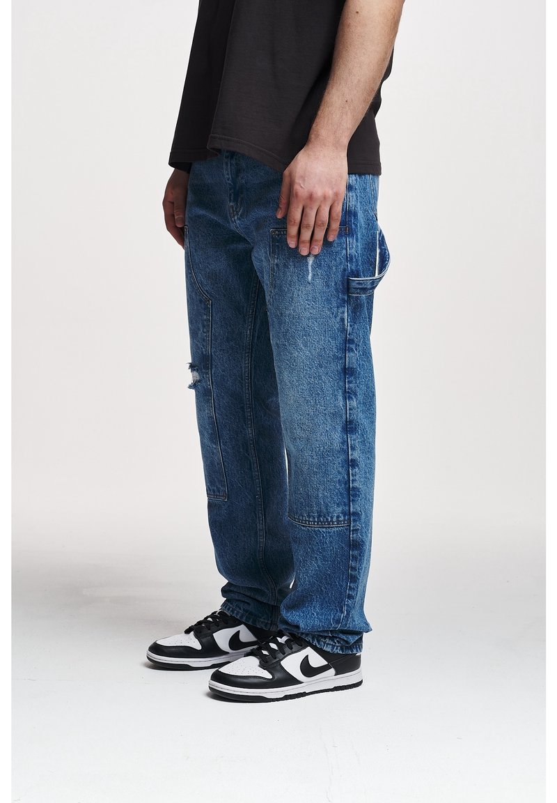 Homme portant un jean denim bleu ample avec des poches latérales et des baskets Nike noires et blanches, debout sur fond blanc.