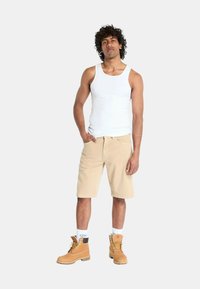 Pantaloni corti beige a ginocchio abbinati a una canotta bianca e stivali da lavoro marrone chiaro. Presenta calze bianche e una postura eretta.