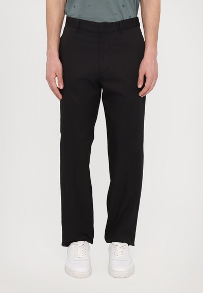 Pantaloni neri realizzati in un tessuto liscio con taglio dritto, dotati di tasche laterali e davanti piatto. Indossati con sneakers bianche.