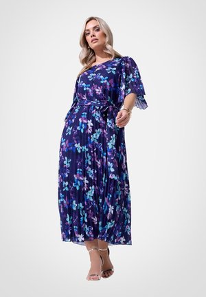 Kobieta w granatowej plisowanej sukience w kwiaty z krótkimi rozkloszowanymi rękawami i srebrnych szpilkach z paskiem wokół kostki na jednolitym tle.