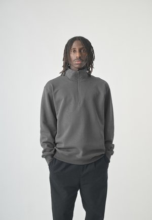 Mann mit Dreadlocks, der einen dunkelgrauen Quarter-Zip-Pullover und schwarze Hose trägt, steht mit den Händen in den Taschen vor einem schlichten Hintergrund.