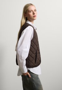 Gilet acolto marrone con colletto, indossato sopra una camicia bianca. Vestibilità casual con tasche laterali e texture liscia. Abbinato a jeans azzurri chiari.