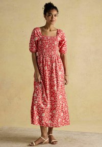 Maxikleid mit rotem Blumenmuster, quadratischem Ausschnitt und kurzen Puffärmeln; verfügt über einen gerafften Oberteil und einen fließenden Rock. Getragen mit hellen Sandalen.