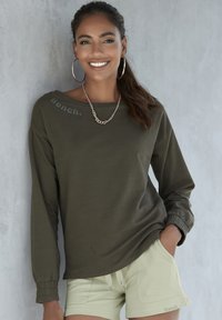 Maglietta a maniche lunghe verde oliva con polsini elasticizzati, con un logo vicino al colletto. Abbinata a pantaloni corti verde chiaro, presentata su uno sfondo neutro.