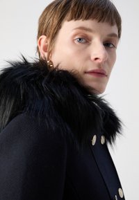 Juodas paltas su dirbtinio kailio apykakle, puošiamas auksiniais mygtukais ir tekstūruotu audiniu. Artimas apykaklės ir modelio veido planas.