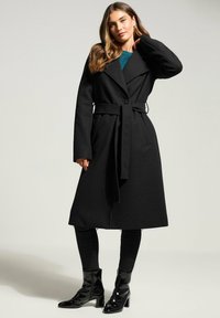 Schwarzer Wollmantel mit maßgeschneidertem Design, weitem Kragen, tailliertem Bund und langen Ärmeln; kombiniert mit schwarzen Jeans und glänzenden Stiefeletten mit Absätzen.