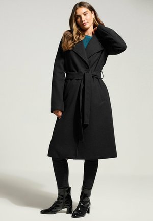 Schwarzer Wollmantel mit maßgeschneidertem Design, weitem Kragen, tailliertem Bund und langen Ärmeln; kombiniert mit schwarzen Jeans und glänzenden Stiefeletten mit Absätzen.