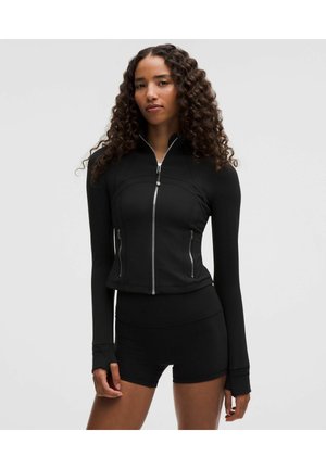 Donna con capelli lunghi e ricci che indossa una giacca nera aderente con zip e pantaloncini neri a vita alta, in piedi davanti a uno sfondo semplice.