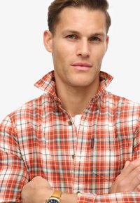 Camicia a quadri arancione, bianca e blu. Presenta una chiusura con bottoni, un colletto a risvolto e una tasca sul petto. Materiale in cotone.