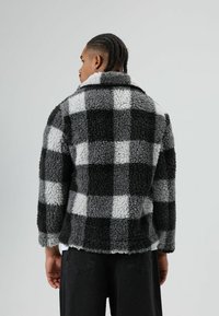 Zwart-grijze geruite fleecejas met kraag, zachte textuur en oversized pasvorm. Knopdetails en ontspannen mouwen zichtbaar.