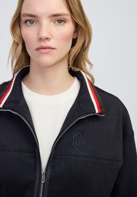 Chaqueta de cremallera en color azul marino, fabricada en tejido suave, con logotipo bordado, cuello a rayas en rojo, blanco y marrón, y un ajuste relajado.