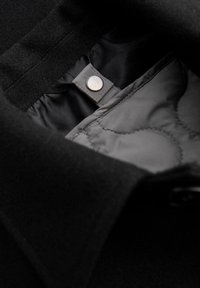 Innenfutter eines schwarzen Wollmantels mit grauer Steppung. Verfügt über einen metallischen Druckknopfverschluss und eine weiche Textur.