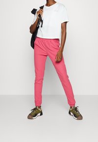 Zkrácené bílé tričko s logem, růžové tepláky s elastickým pasem a hnědé/černé sportovní tenisky se zelenými akcenty.