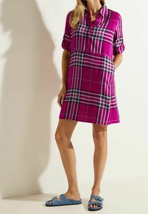 Frau in einem kurzen karrierten Magenta-Kleid mit hochgekrempelten Ärmeln und blauen Sandalen mit Schnallen, die vor einer hellen Wand steht.