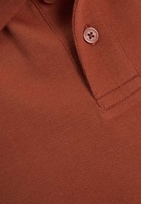 Polo shirt couleur rouille avec un tissu texturé, une patte de boutonnage à deux boutons et un design de col classique.
