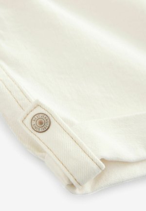 Biely denímový materiál má textúrovanú väzbu s viditeľným detailemi šitia. Na okrúhlej kovovej nálepke je nápis "NEXT JEANS SINCE 1982."