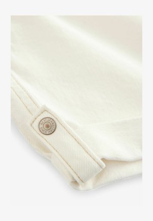 Valge denim kangas on tekstureeritud kudumiga, millel on nähtav õmblusdetail. Ümmargune metalllabel kannab ütlemist "NEXT JEANS SINCE 1982."