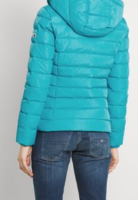 Turquoise gewatteerd jack met een horizontaal doorgestikt patroon, een capuchon en een logo op de mouw. Gecombineerd met een blauwe spijkerbroek en een gladde textuur.