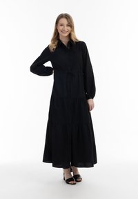Lang sort kjole med knaplukning, lagdelt design og snor i taljen. Stoffet ser let og glat ud. Stylet med sorte sandaler.