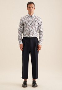 Weißes Hemd mit einem blauen, roten und grünen Blumenmuster; kombiniert mit marineblauen Hosen; schwarze Lederschuhe; taillierte Passform.