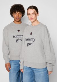 I felpe grigie con collo a giro presentano il logo "tommy girl" in blu navy, con un accento a stella in rosso e bianco. Maniche lunghe con polsini e orlo a costine.