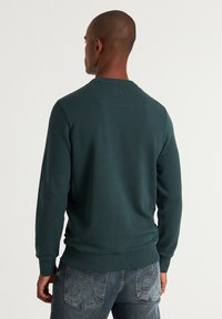 Teal tröja med rund halsringning, ribbade ärmslut och nederkant; har en subtil logotypdetalj på baksidan. I kombination med blekta blå jeans.