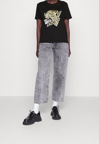 Camiseta de algodón negra con gráfico de leopardo y texto, combinada con pantalones vaqueros de pierna ancha grises con dobladillos deshilachados y zapatos negros de suela gruesa.