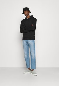 Felpa nera con cappuccio, jeans azzurri chiari, scarpe slip-on grigie. Il modello posa con le braccia incrociate, indossando un cappello a secchiello nero.