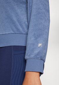 Blaues Velours-Sweatshirt mit gerippten Bündchen. Mit kleinem Logo auf dem linken Ärmel. Kombiniert mit strukturieren Leggings in Marineblau.