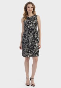 Schwarzes ärmelloses Kleid mit abstraktem weißen Blumenmuster, tailliertem Oberteil und einem ausgestellt Rock. Kombiniert mit schwarzen Riemchen-Heels.