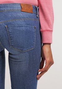 Jeans de denim azul con corte ajustado, con bolsillos traseros y una etiqueta de cuero marrón en la cintura. La tela muestra una ligera textura.