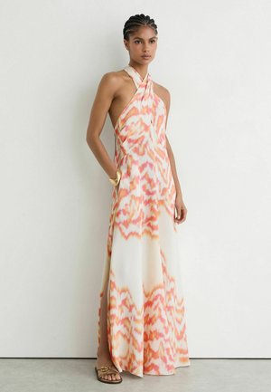 Mujer con cabello trenzado que lleva un vestido largo sin mangas, con cuello halter, con estampado naranja y blanco, con bolsillos laterales y sandalias marrones.