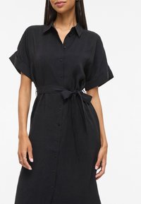 Γυναίκα που φοράει μαύρο φόρεμα με κοντά μανίκια και κουμπιά, με ζώνη στη μέση δεμένη, στέκεται μπροστά σε λευκό φόντο.