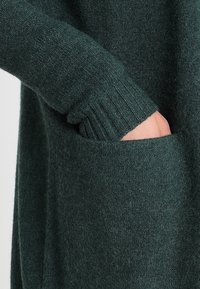 Dark green knitted sweater with a ribbed cuff. The left hand is tucked into a front pocket, showcasing a soft, textured fabric.