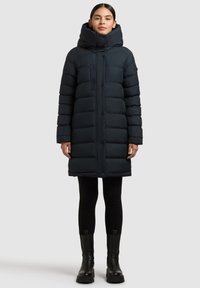 Abrigo largo acolchado en azul marino oscuro, con cuello alto, bolsillo con cremallera y paneles horizontales; combinado con leggings negros y botas gruesas.