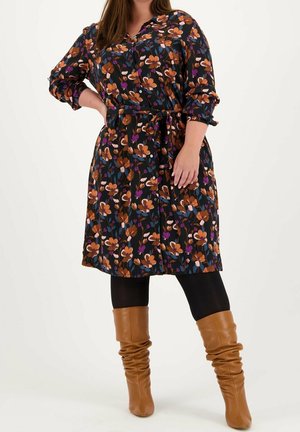 Kobieta ubrana w czarną sukienkę w kwiaty w kolorach pomarańczowym, fioletowym i białym, czarne legginsy oraz beżowe kozaki za kolano, stoi z ręką na biodrze.