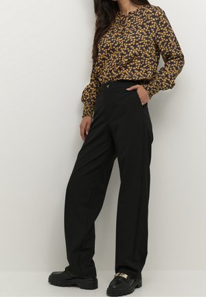 Vrouw draagt zwarte wijde broekspijpen, zwarte loafers en een blouse met lange mouwen met een geel bloemenpatroon op een donkere achtergrond.