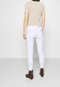 Jeans blancos ajustados con una cintura alta, que cuentan con bolsillos traseros y un detalle de cremallera. Combinados con una blusa beige de manga corta y botas de tobillo marrones.