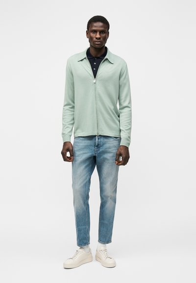 Άνδρας που φοράει ανοιχτοπράσινο ζακέτα με φερμουάρ πάνω από μαύρο πόλο, μπλε τζιν, λευκές κάλτσες και λευκά αθλητικά, στέκεται μπροστά σε λευκό φόντο.