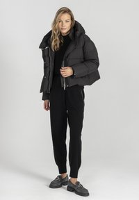 Krótka czarna kurtka puffer z kapturem, błyszczącymi zamkami błyskawicznymi i prostokątnym krojem. Połączona z czarnymi dzianinowymi spodniami i ciemnym obuwiem.