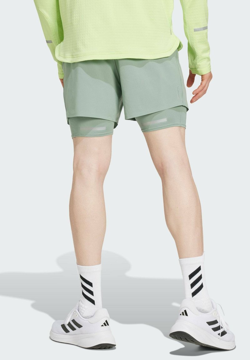 Beine einer Person, die hellgrüne, geschichtete Shorts, weiße Adidas Turnschuhe mit schwarzen Streifen und weiße Kniestrümpfe mit schwarzen Streifen trägt.