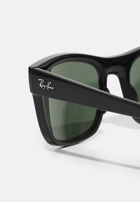 Ochelari de soare Ray-Ban negri, cu lentile mari verzi, un cadru pătrat îndrăzneț și un finisaj lucios, având logo-ul brandului pe lateral.