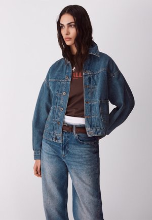 Vrouw die een donkerblauwe spijkerjack en spijkerbroek draagt, een bruin T-shirt met grafische print en een riem, staand met één hand in de zak en zijwaarts kijkend.