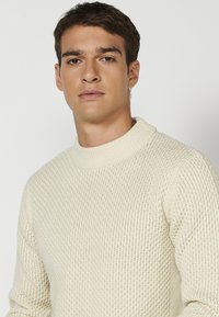 Crème-kleurige gebreide trui met een hoge halslijn en een textuurpatroon. De stof lijkt dik en comfortabel te zijn met ribbelaccenten op de kraag.