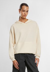 Krémově zbarvená oversized mikina s kapucí a přední kapsou, stahovací šňůrou a žebrovanými manžetami, doplněná o tmavě šedé kalhoty. Hladká textura.