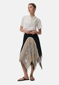 Witte gedrapeerde top gecombineerd met een geplooide zwarte rok met beige en zwarte kantpatronen en een uitlopende zoom. Zwarte teenslippers maken de look compleet.